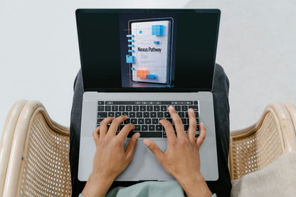 Top-down view of a person typing on a silver laptop while sitting in a woven chair. The laptop screen displays a digital course or document titled 'Nexus Pathway'.